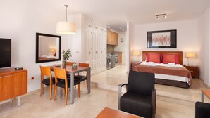 Habitación de hotel con cama, mesitas de noche, escritorio y sillas, televisor, zona de comedor con mesa y sillas, y cocina.