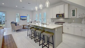 A modern kitchen with white cabinets, a central island with bar stools, a microwave, and a TV mounted on the wall. - Pecan Pie 236 BR (Port Aransas)