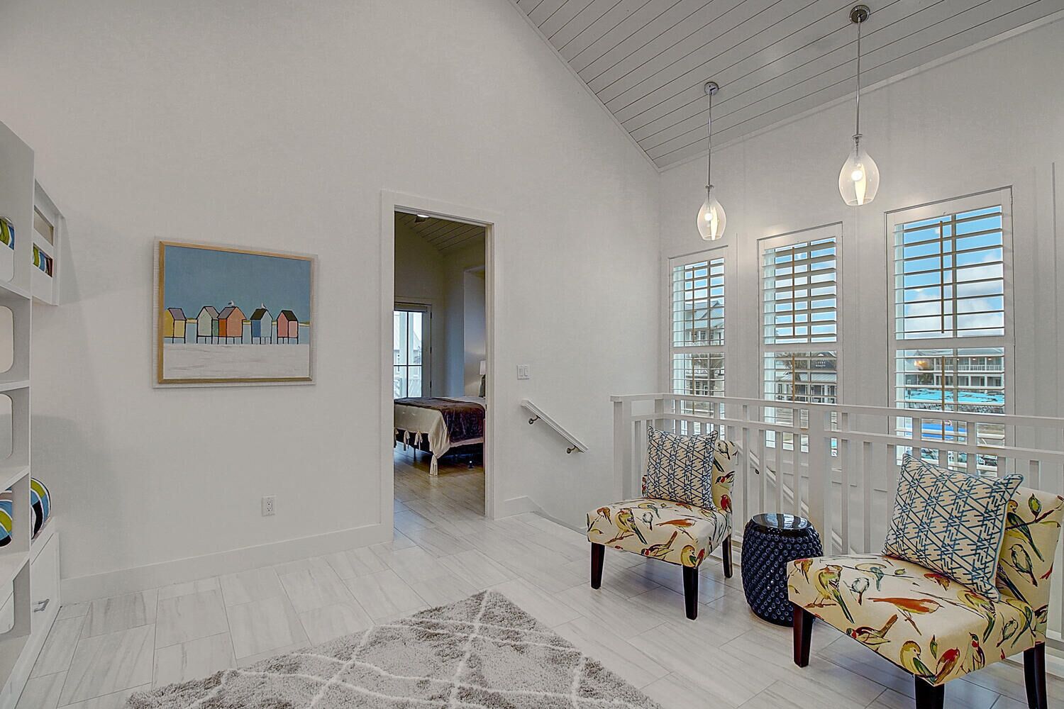 A spacious living area with a high ceiling, a staircase leading to an upper floor, and large windows with blinds.