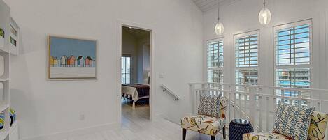 A spacious living area with a high ceiling, a staircase leading to an upper floor, and large windows with blinds.