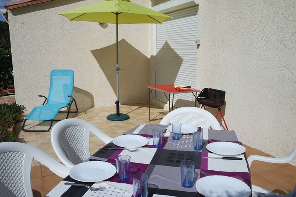 Eine Außenpatio mit einem für vier Personen gedeckten Tisch, umgeben von weißen Stühlen, einem gelben Sonnenschirm und einem Grill.