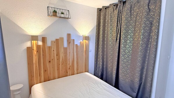 Un dormitorio con cabecera de madera, cama blanca, cortinas estampadas y una estantería con plantas.