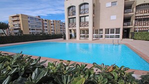 A swimming pool surrounded by a building with balconies and a hedge in the foreground.