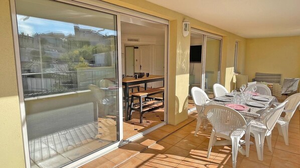 Un patio soleado con una mesa y sillas de comedor, una puerta corrediza de vidrio que da a un balcón y una pequeña zona de cocina.