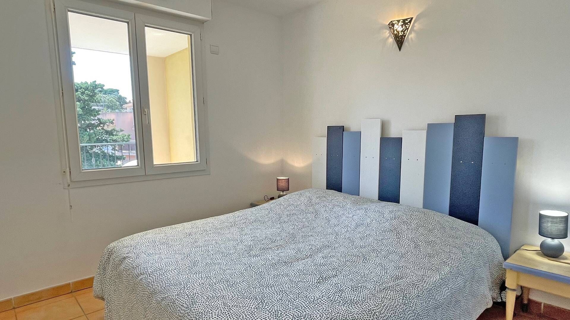 Un dormitorio con cama, ventana, lámpara de pared y mesita de noche con lámpara.