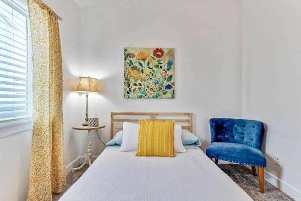 A bedroom with a bed, a blue chair, a lamp, a painting, and a window with curtains.