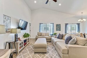 A spacious living room with a sofa, ottoman, and a flat-screen TV mounted on the wall.