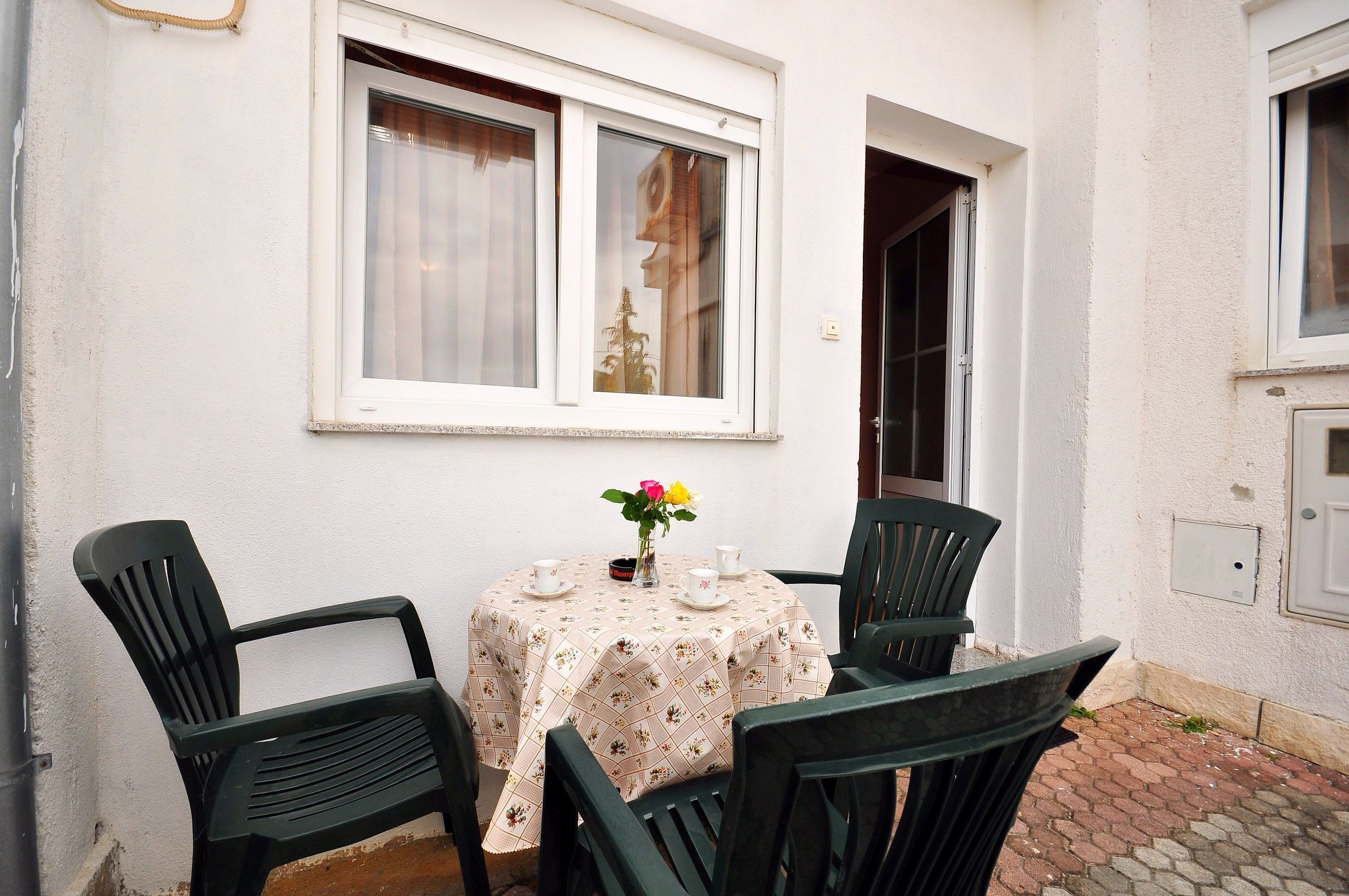 A small outdoor seating area with a table and chairs, a window with curtains, and a door leading to another room.