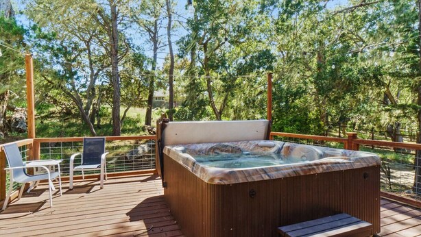A hot tub on a wooden deck with chairs and a bench, surrounded by trees.