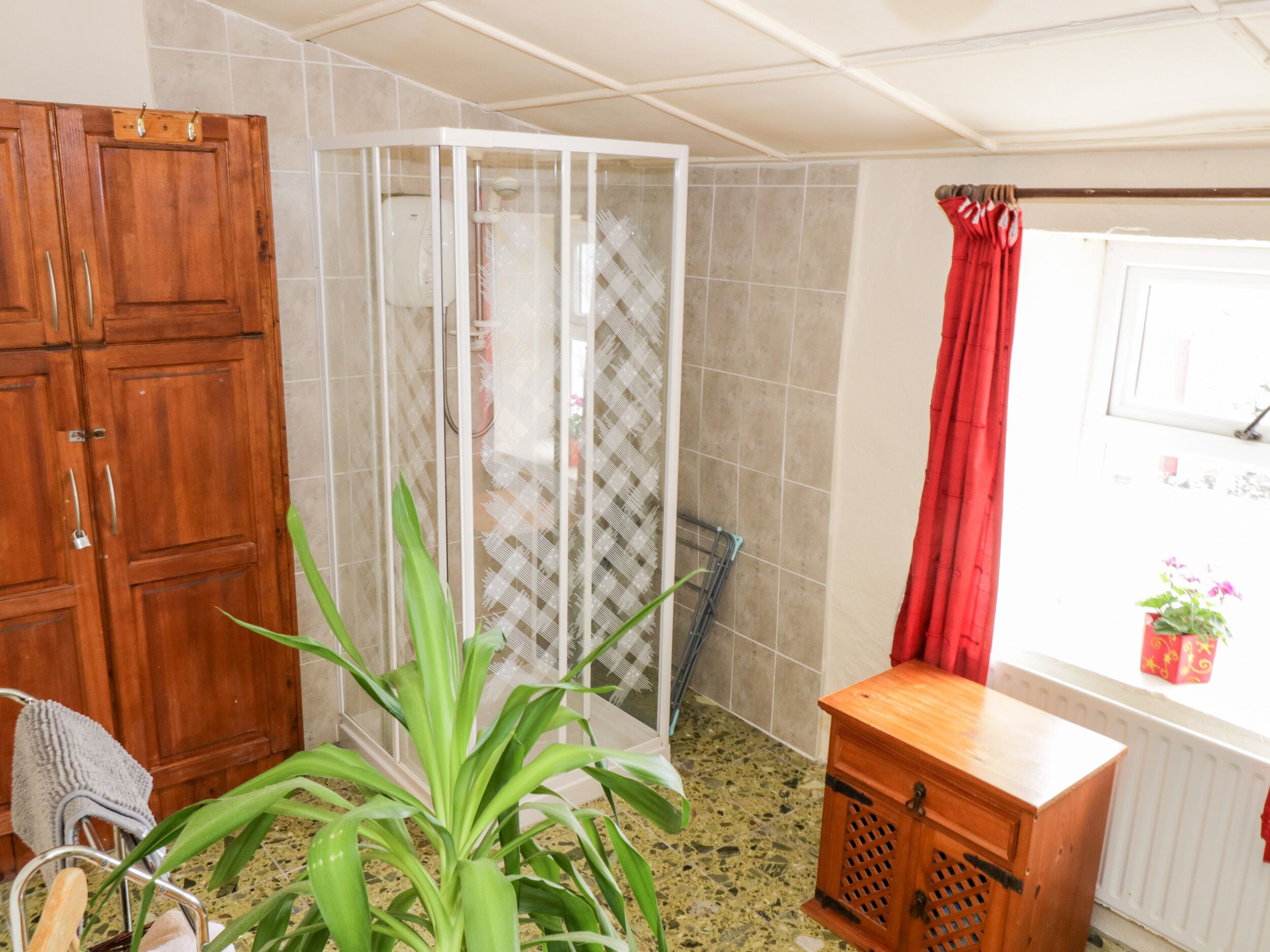 A bathroom with a shower, a wooden cabinet, a plant, and a window with a red curtain.