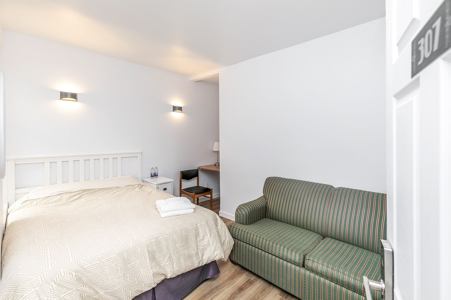 Un dormitorio con una cama, un sofá a rayas verdes, una mesita de noche y dos luces fijadas a la pared.