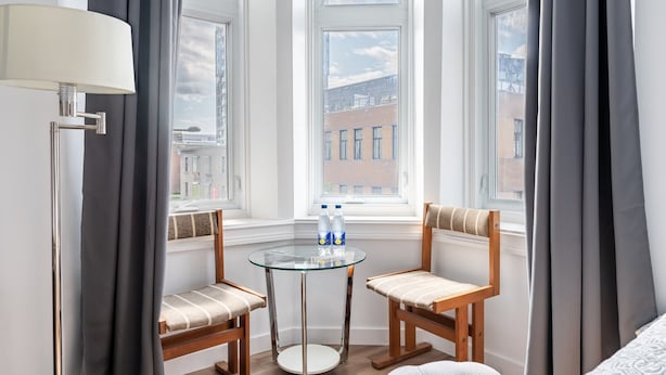A hotel room with a glass table, two wooden chairs, a lamp, and a view of buildings through large windows. - Hotel le Coach (Montreal)