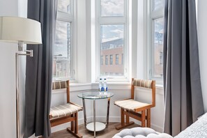 A hotel room with a glass table, two wooden chairs, a lamp, and a view of buildings through large windows. - Hotel le Coach (Montreal)