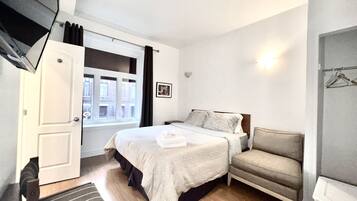 Un dormitorio con cama, mesita de noche, silla, ventana y puerta.