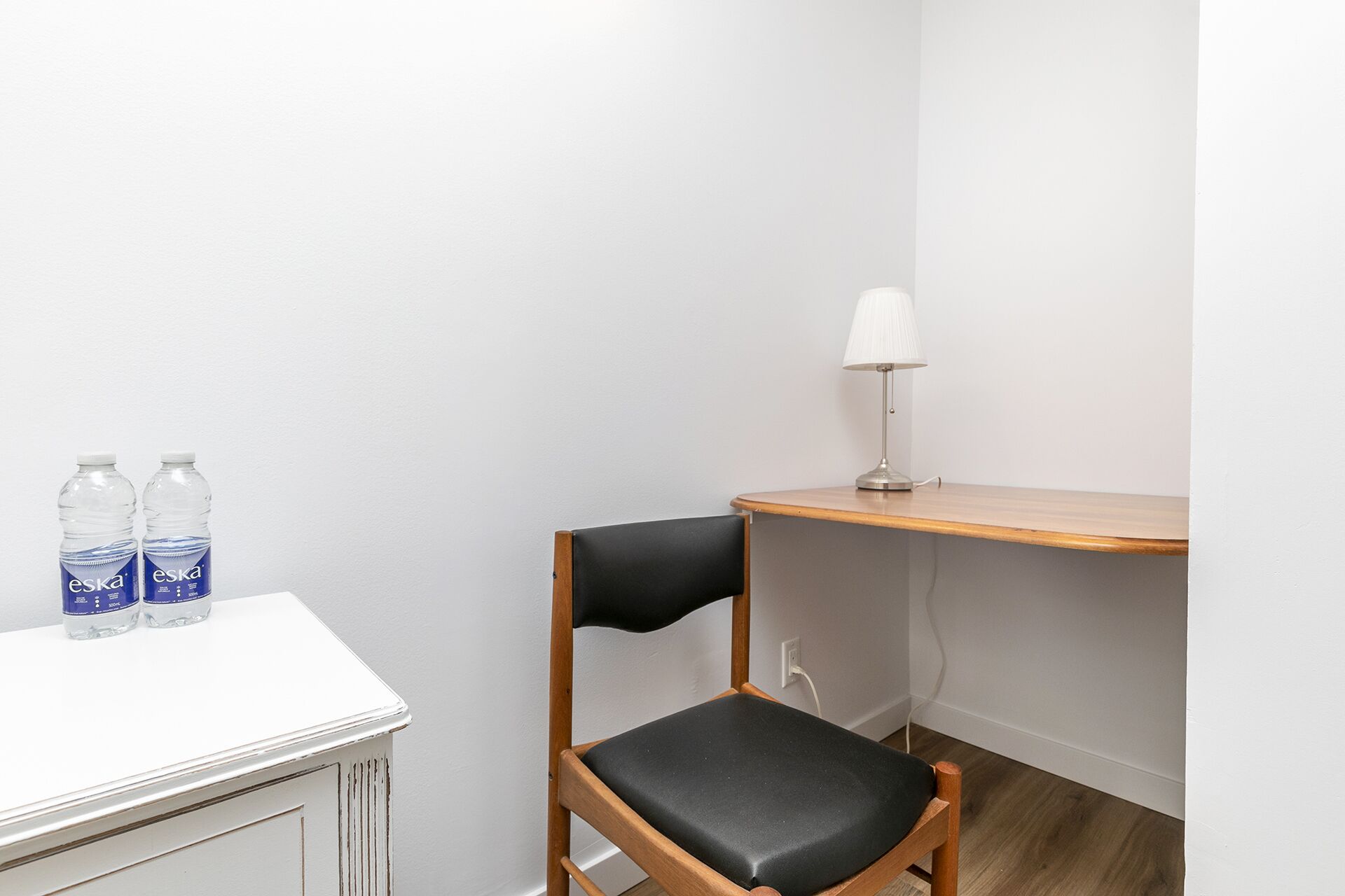 A small, clean room with a wooden chair, a white desk, and two bottles of water.