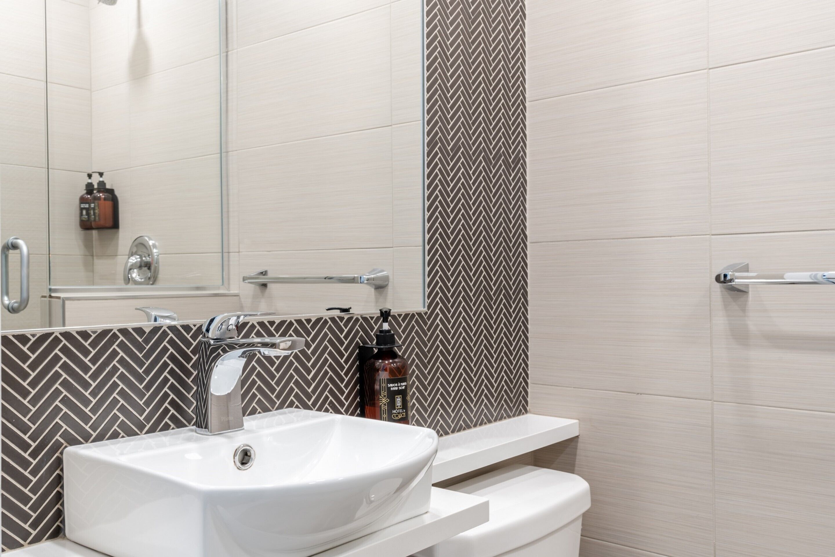 A modern bathroom with a white sink, chrome faucet, and a mirror above it. There is a shower area with a glass door and a towel rack.