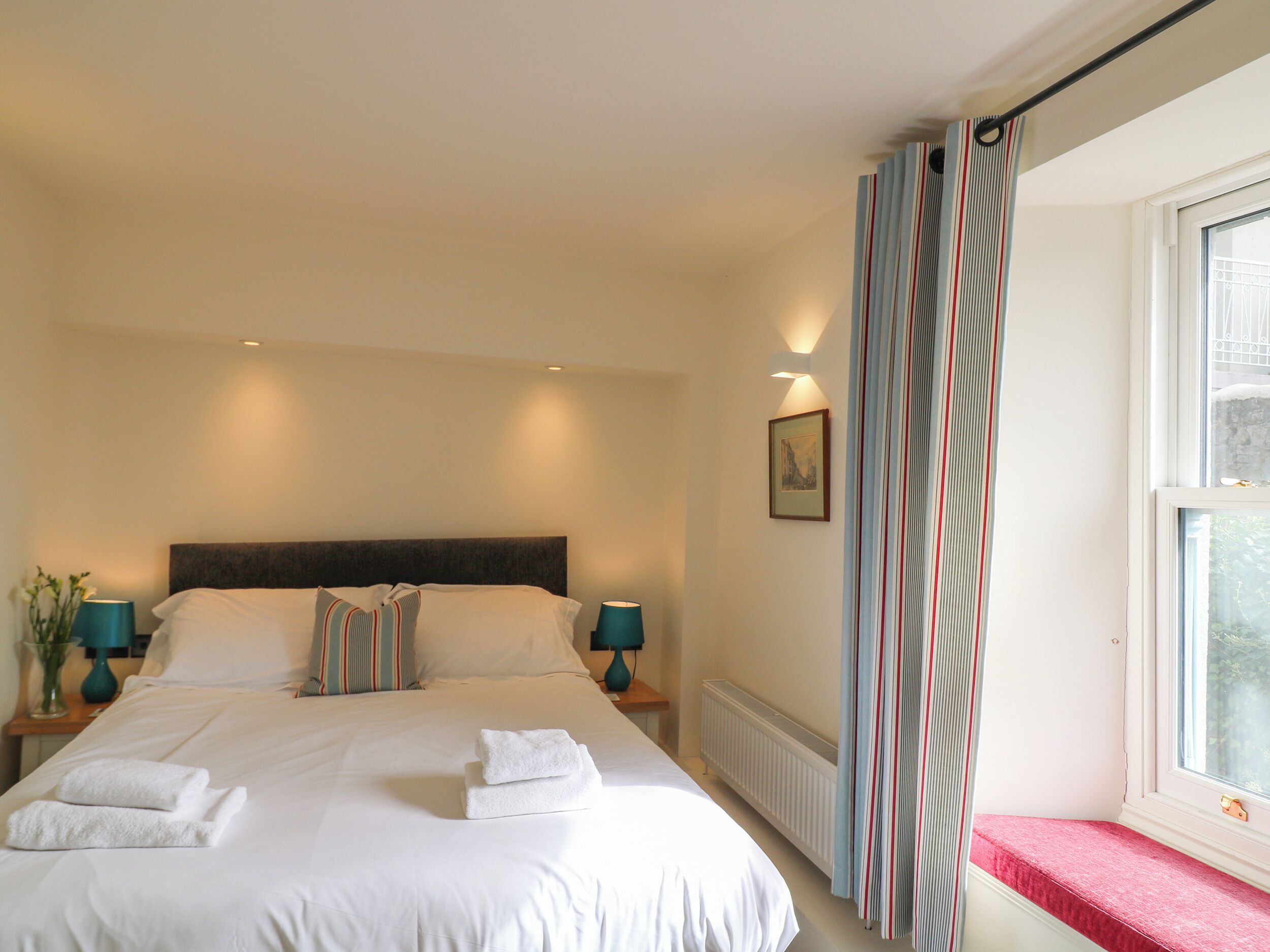 Una habitación de hotel con una cama grande, dos mesitas de noche con lámparas, un radiador, una ventana con cortinas y un cuadro en la pared.