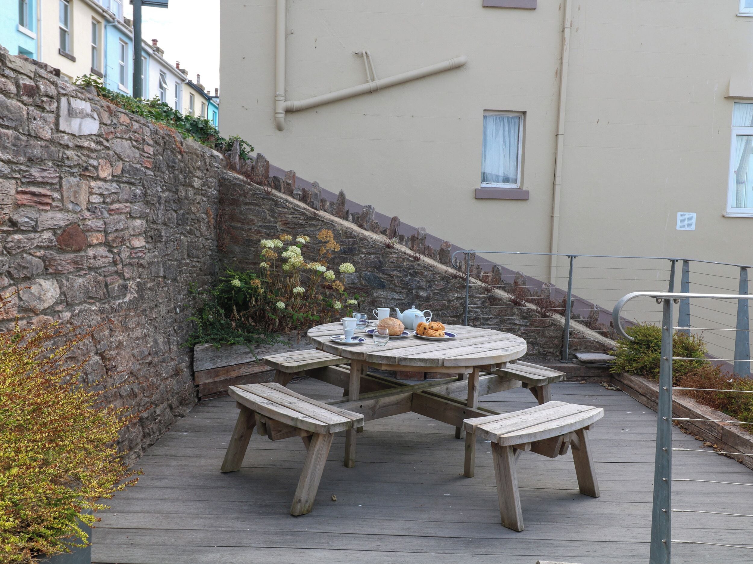 Une table ronde en bois, entourée de bancs, un mur de pierre et, au fond, un bâtiment avec des fenêtres.