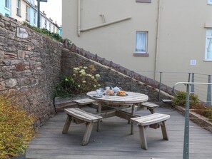 Una mesa redonda de madera con bancos, un muro de piedra y un edificio con ventanas al fondo.