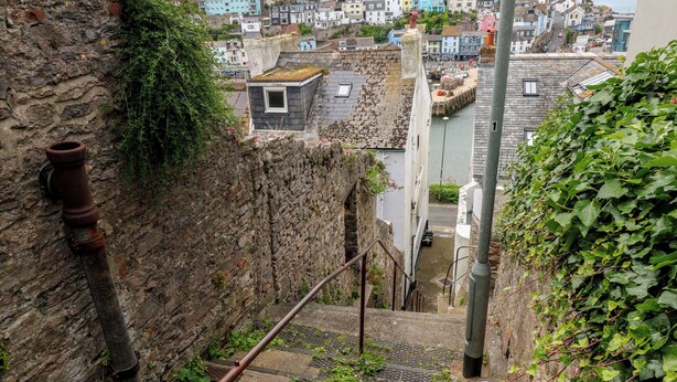 Eine enge Treppe mit Metallhandlauf, einer Steinwand zur Seite und Blick auf eine Stadt mit bunten Gebäuden im Hintergrund.