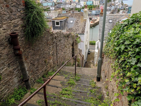 Una escalera estrecha con barandilla de metal, pared de piedra a un lado y vista de un pueblo con edificios de colores al fondo.