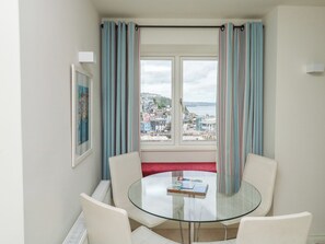 A room with a round glass table, white chairs, a window with striped curtains, and a view of a coastal town.