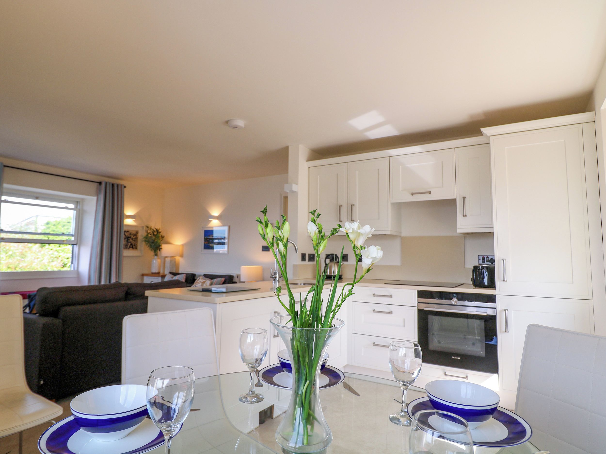 Une cuisine moderne avec des meubles blancs, une table à manger avec de l’assiette bleue et blanche, et un vase rempli de fleurs.