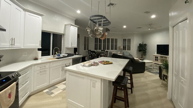 A modern kitchen with white cabinetry, a large island, and a dining area with a sofa and TV. - RELAXING OASIS-Private Lanai Heated POOL & SPA.  4 Bed, 3.5 Bath / Beach 10 min (Bonita Springs)