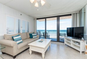A living room with a beige sofa, a white coffee table, a flat-screen TV, and a balcony with a view of the ocean.