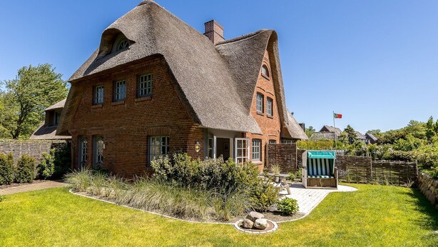 Una casa grande de ladrillo con techo de paja empinado, una estructura de madera adosada y un jardín bien cuidado.