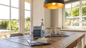Una mesa de madera con una botella, vasos y un menú en una habitación luminosa con ventanas amplias.
