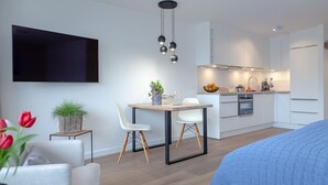 Ein modernes, kompaktes Wohnzimmer mit Essbereich, Küchenzeile und einem an der Wand befestigten Flachbildfernseher.