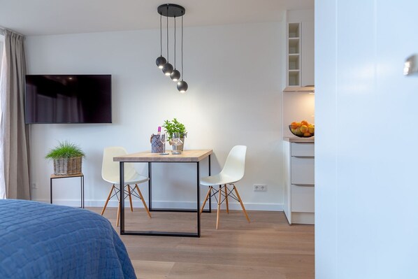 A modern hotel room with a wooden dining table, two white chairs, a TV mounted on the wall, and a kitchen area with a fruit bowl.