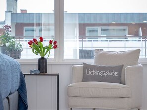 A room with a view of a building through a window, a chair with a pillow, and a vase with red flowers on a side table.