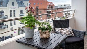 Ein Balkon mit Korbmöbeln, einer Topfpflanze und Blick auf Stadtgebäude.