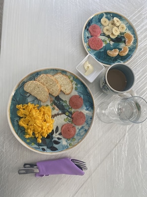 Mesa posta para o café da manhã com ovos mexidos, fatias de linguiça, torradas, bananas e uma xícara de café.