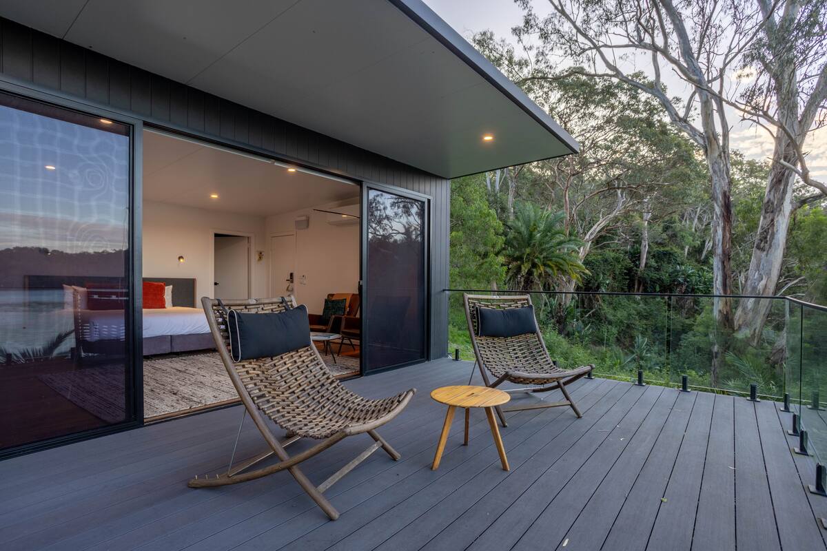 A modern outdoor deck with wicker chairs and a small table, overlooking a lush garden and a bedroom with a large window.