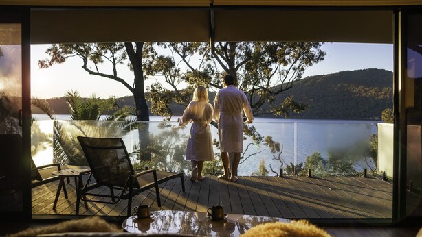 A couple standing on a wooden deck overlooking a lake, with a chair and table in the foreground. - Marramarra Lodge (Berowra Waters)