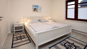 A white bedroom with a bed, bedside tables, a window with blinds, and a rug with a ship's wheel design.