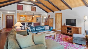 Ein geräumiges Wohnzimmer mit hohen Decken, Holzbalken, einem Ledersofa, einem Fernseher und einer Küche im Hintergrund.