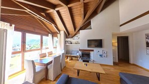 A spacious living area with wooden beams, a fireplace, a television, and a view of the outdoors.