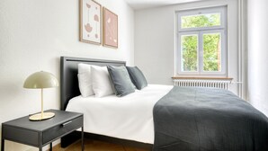 Et soveværelse med seng, natbord, lampe, indrammet kunst og et vindue med udsigt til grønne omgivelser.