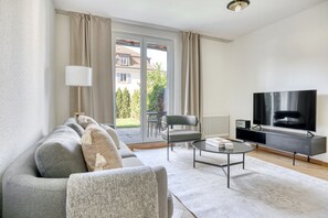 Un salon moderne avec un canapé gris, une table basse ronde, un téléviseur posé sur un meuble et une vue sur un jardin, visible à travers une porte-fenêtre coulissante.