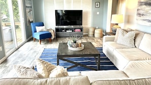A living room with a flat-screen TV, a blue armchair, a beige sofa, a coffee table, and a large window with blinds. - Waterfront Bethany Beach Cottage, Walk to Beach, Fish, Kayak - Dewey Rehoboth (Bethany Beach)