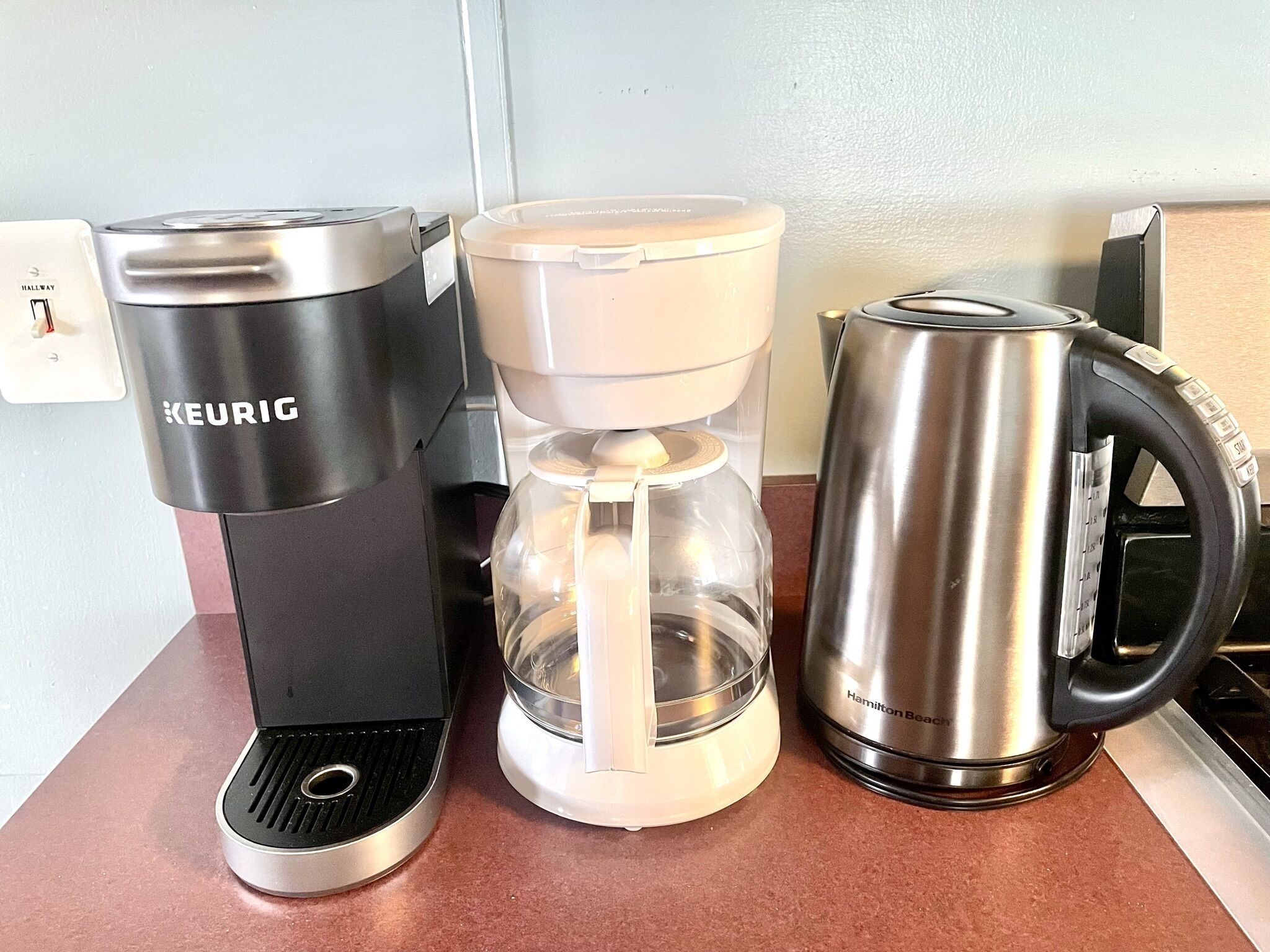 A Keurig coffee maker, a glass carafe, and a Hamilton Beach electric kettle on a countertop.