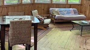 A wooden interior with a dining table, chairs, a sofa, and large windows offering a view of greenery. - Island Tree House Glamping (Monmouth)