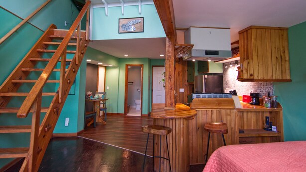 A spacious room with a wooden staircase, a kitchen area with bar stools, and a bed with a pink blanket.