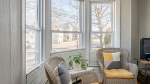 Una habitación acogedora con ventanas amplias, sillones de mimbre, una mesita con una planta y un televisor sobre un mueble de madera.