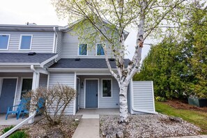 A two-story residential building with a front porch, a tree, and a small garden area.