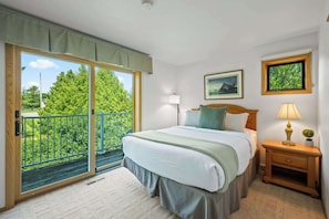 A bedroom with a bed, a nightstand, a lamp, a picture frame, and a balcony with a view of greenery.